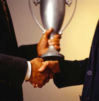ein Handschlag zweier Männer ist zu sehen, während einer von ihnen einen Pokal in der anderen Hand hält 
