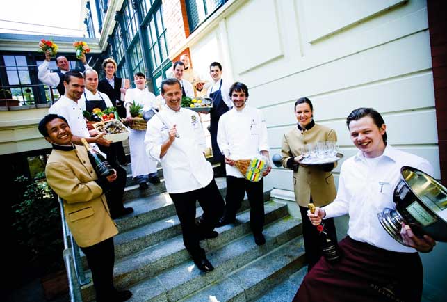 ein Hotelteam auf den Treppen, alle lächelnd mit Blumen und Champagnerflaschen und Serranoschinken 