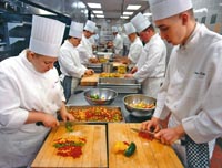 mindestens sechs Köche sind zu sehen, aufgereiht wie sie Gemüse auf Schneidbrettern für die Gerichte kleinschneiden 