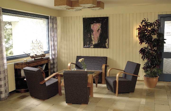 Hotellobby mit Polstermöbeln, und einem großen Fenster, schlicht gehalten und einer großen Topfpflanze 