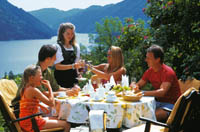 ein Tisch mit Gästen im freien mit Blick auf den See und Berge wird von einer Kellnerin bedient, alle sehen zufrieden und fröhlich aus 