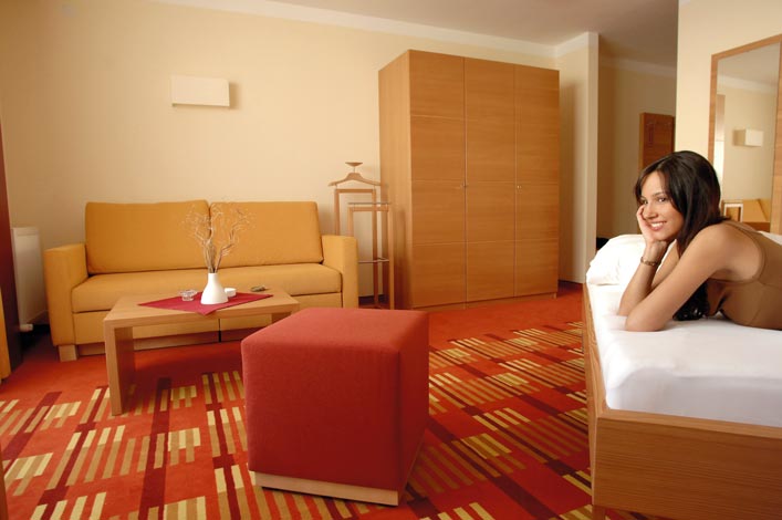 eine Dame liegt auf dem Bett ihres Hotelzimmers mit dem Arm unter ihrem Kopf, des weiteren sieht man einen Kleiderschrank ein kleines Sofa als auch einen Hocker, das Zimmer wird in den Farben Gelb und Rot gehalten 