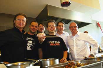 einige Herren versammeln sich hinter dem Herd, der Mann in der Mitte hält ein Messer in der Hand 
