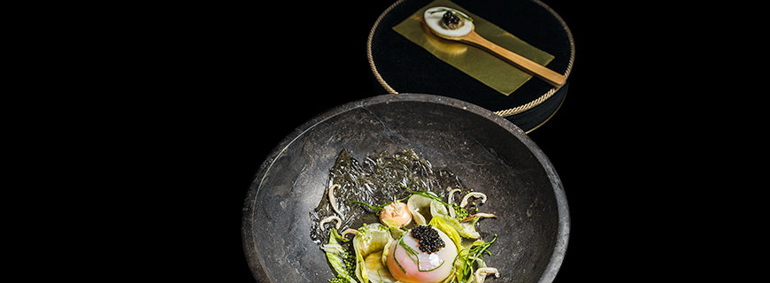 Joachim Wisslers Gericht „Landei Miso gebeizt, Lachscreme, Eisbergsalat & d’Aquitaine-Kaviar“ in einem anthrazitfarbenen tiefen Teller angerichtet. Dazu wird ein Kräcker mit Kaviar gereicht.