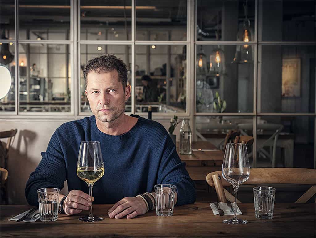 Til Schweiger sitzt mit einem vollen Weinglas am Tisch