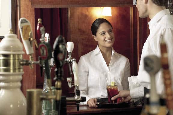 eine Frau und ein Mann sehen sich an, während er ihr den Cocktail aufs Tablett stellt 