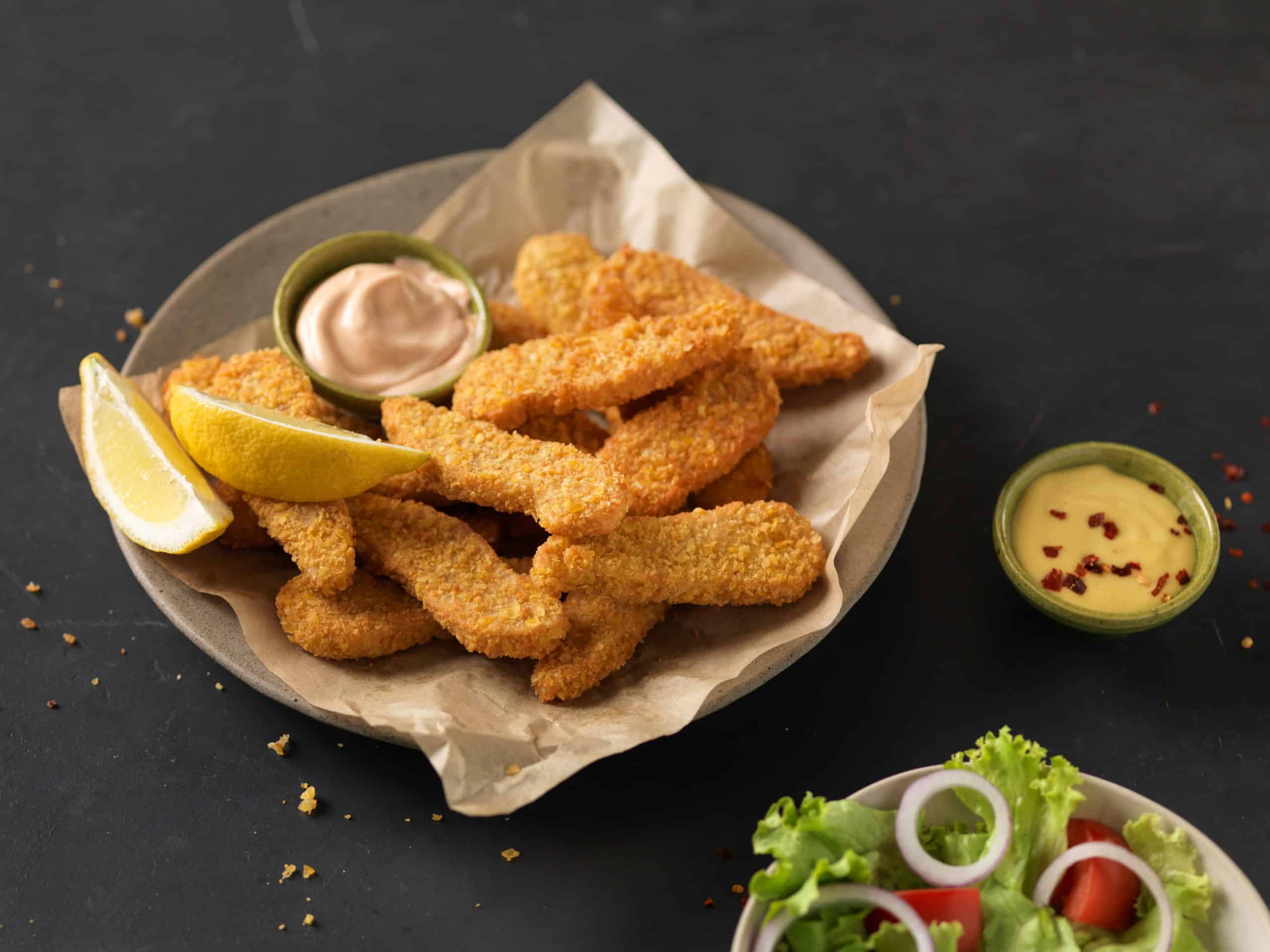 Pflanzliche Crispy Tenders von THE GREEN MOUNTAIN mit goldbrauner Panade, Zitronenspalten und zwei Dips.