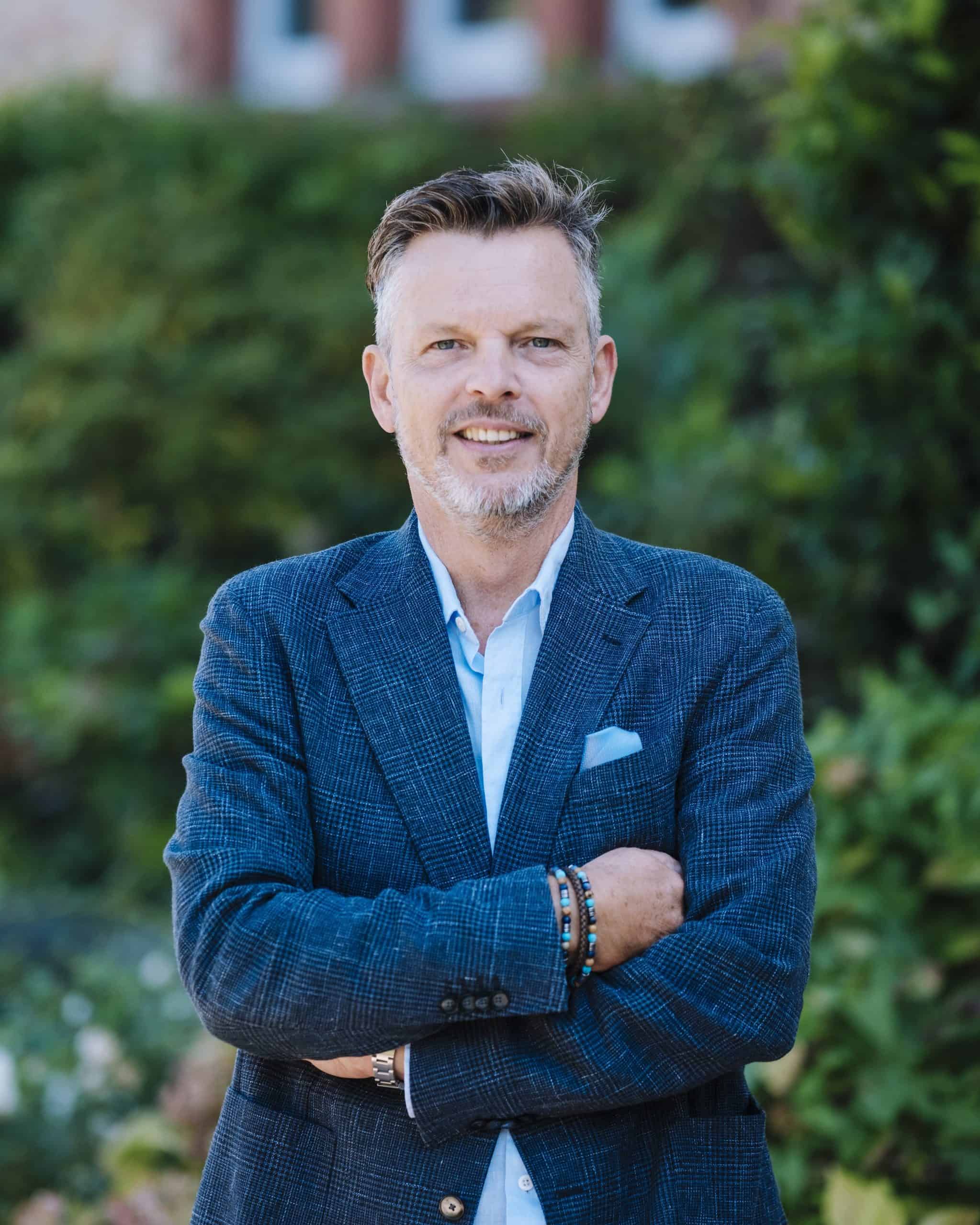 Michael Schorm, Gründer von SCHORM – Hospitality Experts, im blauen Sakko mit verschränkten Armen vor grünem Hintergrund.