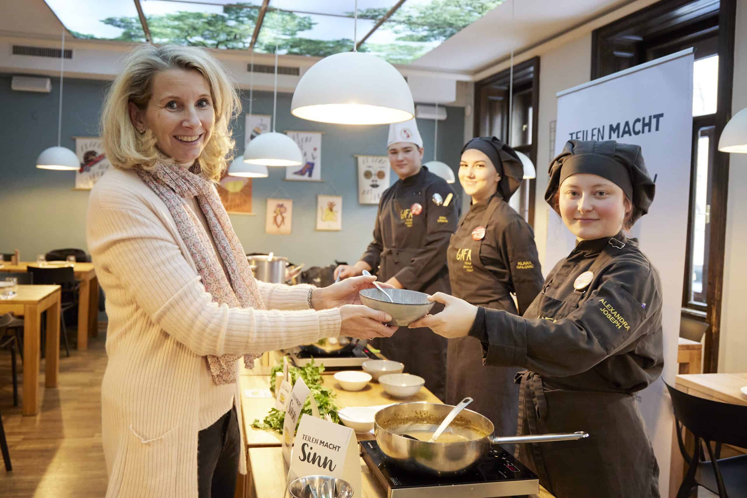 Geschäftsführerin Alexandra Gruber von Die Tafel Österreich nimmt bei „Suppe mit Sinn“ eine Suppenschale von einer Köchin entgegen. Daneben stehen eine weitere Köchin und ein Koch.