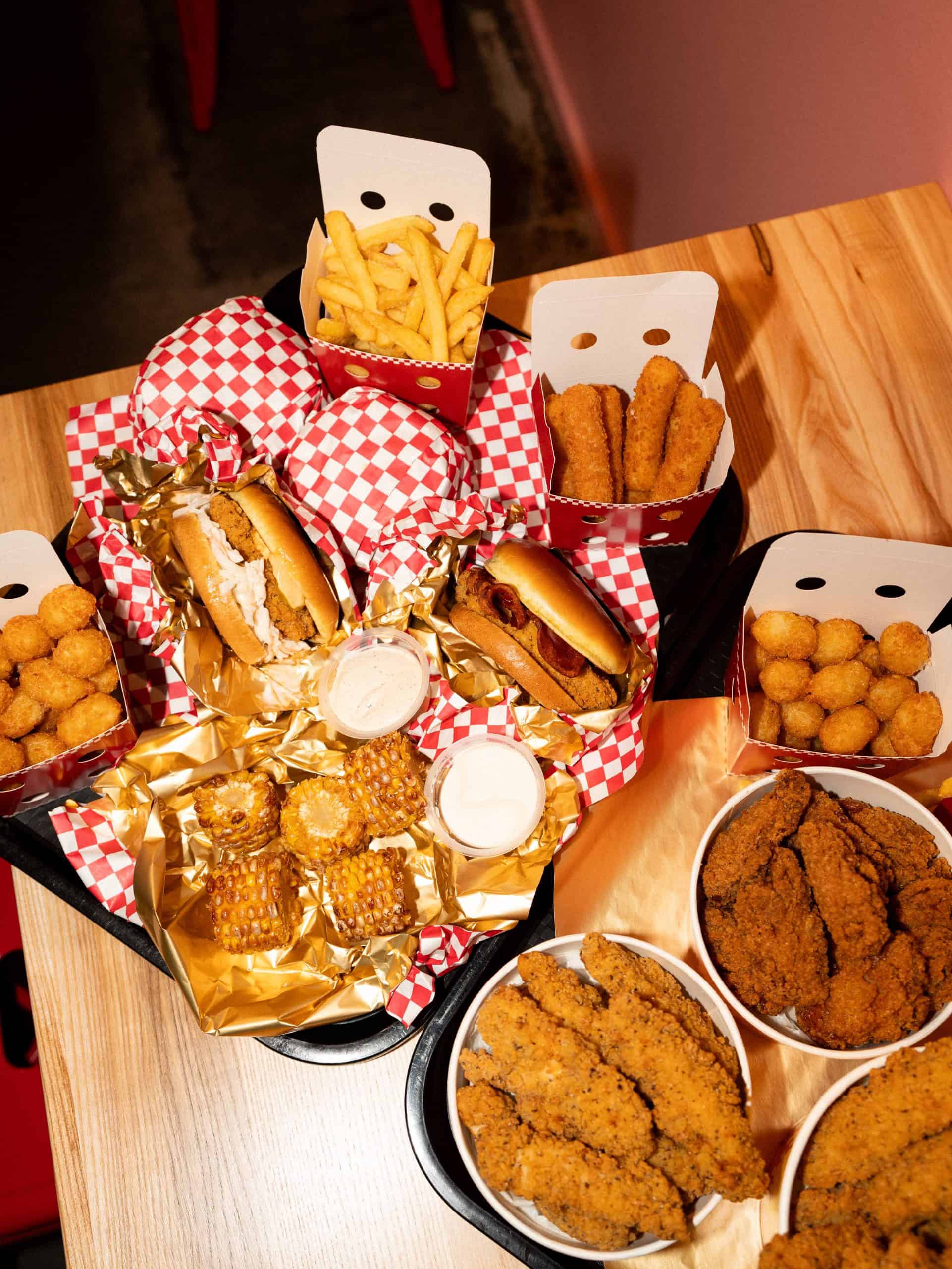 Burger, panierte Hähnchenstücke, Pommes, Mais und Dips von Loco Chicken auf einem Holztisch.